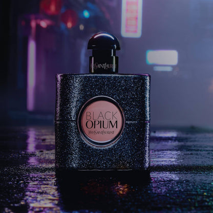 YSL Black Opium perfume bottle on a reflective surface with blurred lights in the background
