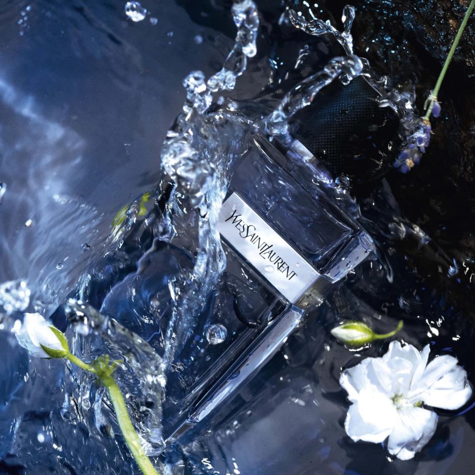 Yves Saint Laurent Y EDP perfume bottle with water splashes and flowers in a dark setting
